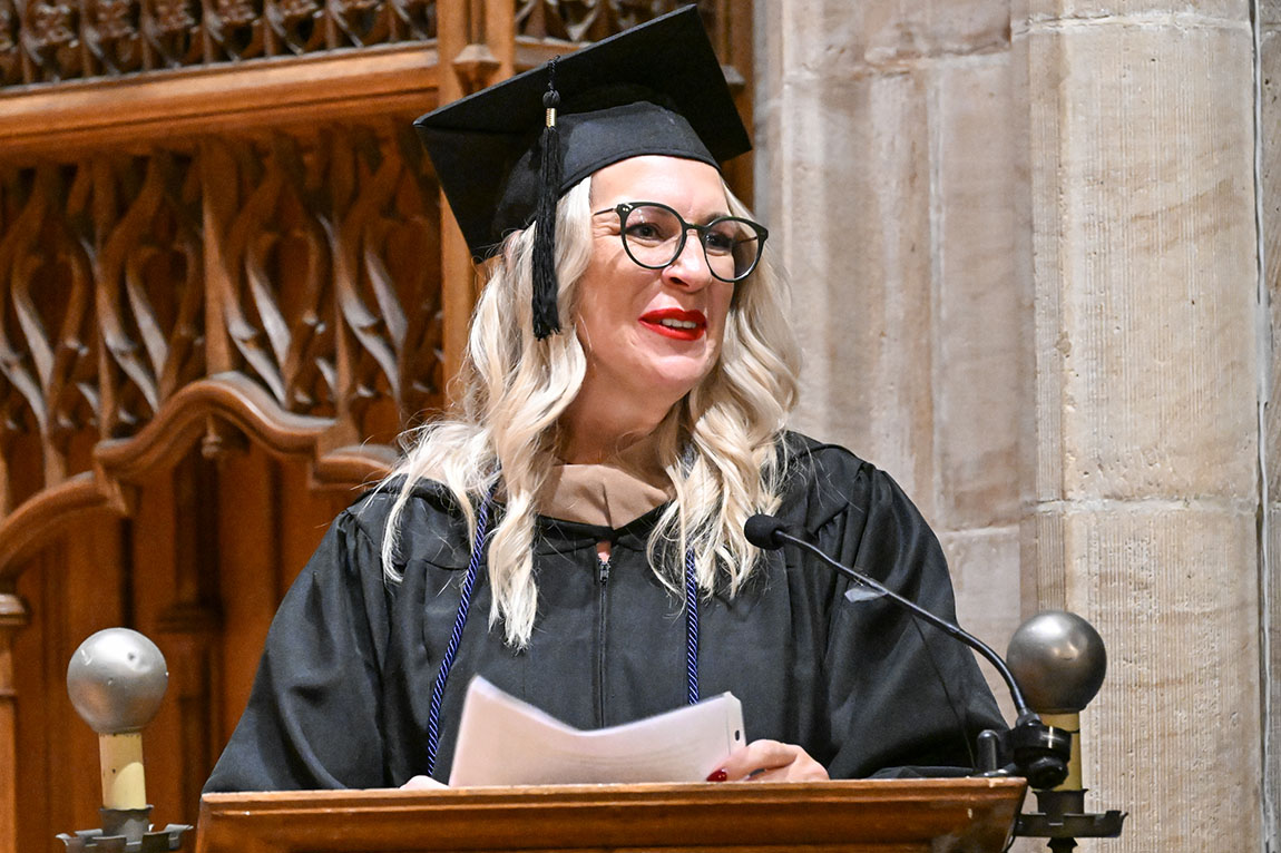 A graduate in cap and gown and glasses with long blond hair speaks at a podium